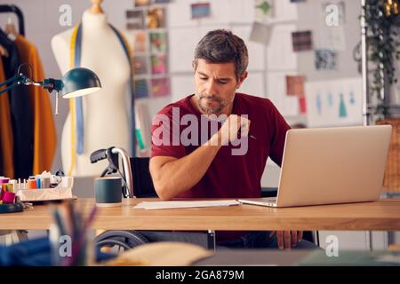 Reifer Männlicher Modedesigner Im Rollstuhl Sitzt Am Schreibtisch Im Studio Und Arbeitet Am Laptop Stockfoto