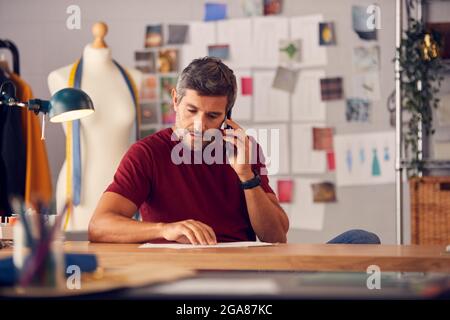 Reifer Männlicher Modedesigner Im Studio, Der Am Schreibtisch An Skizzen Oder Dokumenten Arbeitet Und Telefongespräche Führt Stockfoto