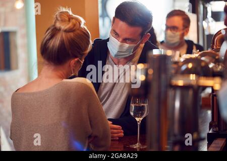 Das Paar Trägt Masken Am Date, Das Während Der Gesundheitpandemie Am Counter Der Geschäftigen Bar Sitzt Stockfoto