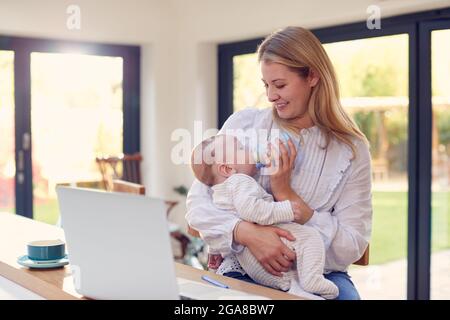 Berufstätigen Mutter Mit Laptop Zu Hause, Während Fütterung Baby Sohn Auf Knie Mit Flasche Stockfoto