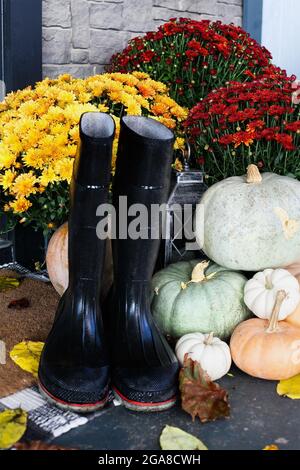 Regenstiefel sitzen auf der Türmatte der Veranda, die für den Herbst mit weißen, orangefarbenen und grauen Kürbissen und Müttern dekoriert wurde. Selektiver FOC Stockfoto