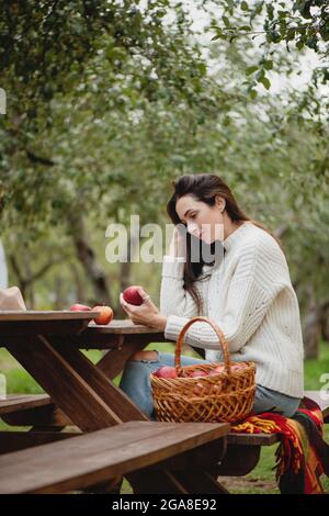 Schöne Frau, die im Apfelgarten mit Eimer voller roter Äpfel auf dem Tisch sitzt. Stockfoto