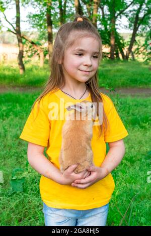 Kleines nettes Mädchen spielt mit einem echten Ingwer-Kaninchen auf einem Hintergrund von grünen Pflanzen. Im Sommer Aktivitäten im Freien für Kinder mit Haustieren. Mädchen spielt mit Stockfoto