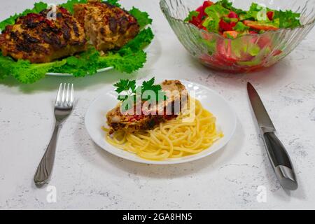 Pasta mit einem Stück Schweinefleisch, das im Ofen gekocht und vor dem Hintergrund einer Salatschüssel im Ofen gebacken wurde, und einem großen Stück gebackenem Fleisch auf einem Teller Stockfoto