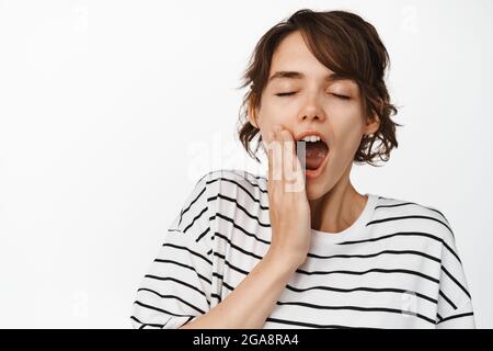 Nahaufnahme einer müden jungen Frau, die gähnt, Müdigkeit empfindet, ein schlaffes Gesicht am frühen Morgen, das in gestreiftem T-Shirt auf weißem Hintergrund steht Stockfoto