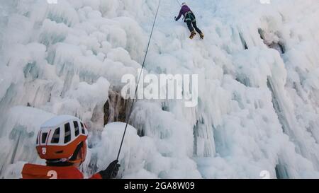 Mann steht und kontrolliert ein Sicherheits-Top-Seil, während Weibchen mit Eiskletterausrüstung auf einem gefrorenen Wasserfall klettert Stockfoto