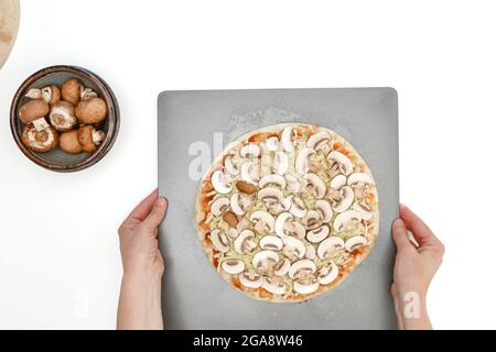 Rohe Pizza mit Tomatensauce, Mozzarella-Käse und Crimini-Pilzen aus nächster Nähe auf der Backform, flaches Lay, weißer Hintergrund, Frauenhände Stockfoto