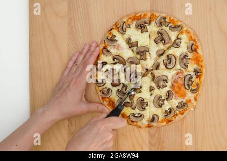 Frau schneidet Pilzpizza mit einem Pizzaschneider, Nahaufnahme von oben, Holzhintergrund Stockfoto