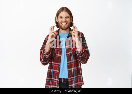 Hoffnungsvoller glücklicher Kerl kreuzt die Finger für viel Glück, lächelt aufgeregt, wartet auf Traum wahr werden, Wunsch, vor weißem Hintergrund stehen Stockfoto