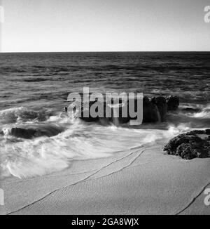 Schwarz-weiß-Schuss von Meereswellen, die auf die Felsen am Strand krachen Stockfoto