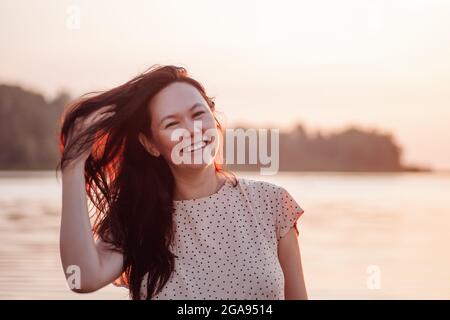 Lächelnde Frau im Freien. Nahaufnahme des Porträts einer glücklichen asiatischen Frau vor dem Hintergrund von Strand und Meer, die mit langen dunklen Haaren spielt und den Sonnenuntergang genießt Stockfoto
