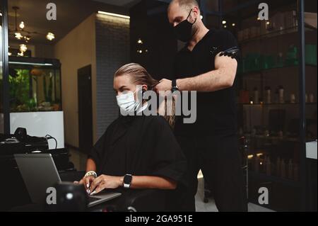 Gutaussehender professioneller Friseur in schwarzem Outfit kämmen Haare von Geschäftsfrau, Kunde besucht Schönheitssalon und arbeiten Remote auf Laptop Stockfoto