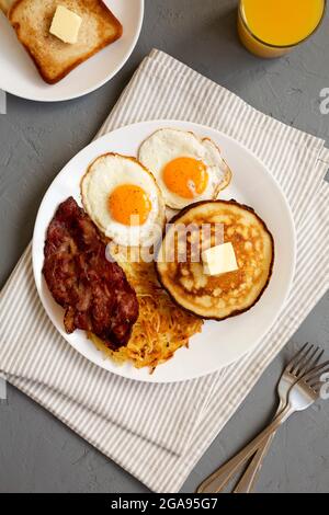 Komplettes amerikanisches Frühstück mit Speck, Hash Browns, Eiern und Pfannkuchen auf einem Teller auf grauem Hintergrund, Draufsicht. Stockfoto