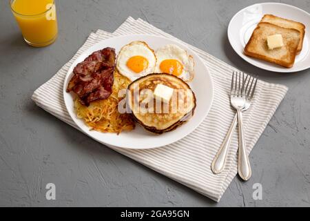 Komplettes amerikanisches Frühstück mit Speck, Hash Browns, Eiern und Pfannkuchen auf einem Teller auf grauem Hintergrund, Seitenansicht. Stockfoto