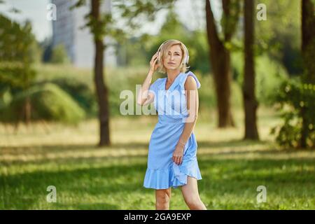 Seriöse blonde Frau mit kabellosen Kopfhörern geht im Park. Stockfoto