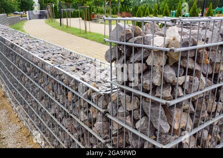 Eine lange Mauer aus Gabionen, die die belebte Straße vom Parkgelände trennt. Gartenarchitektur. Stockfoto