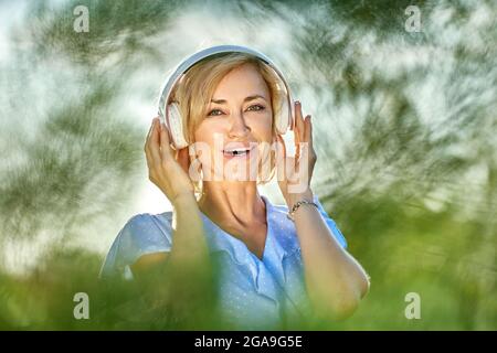 Lächelnde blonde Frau mit schnurlosen Kopfhörern geht im Park. Stockfoto