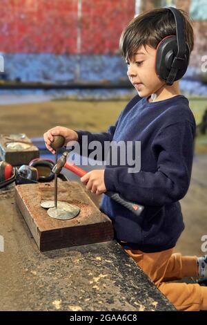 Brünett kleiner Junge mit einem Kopfhörer hält einen Hammer in einem Holzbrett Tisch hämmern Zinnherzen, andere Werkzeuge im Hintergrund. Vertikal, verschwommen Stockfoto