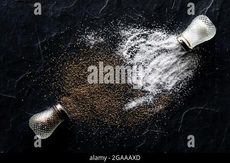 Draufsicht auf Salz- und Pfefferstreuer, die mit Salz und Pfeffer auf schwarzen Schiefer übergossen wurden. Stockfoto