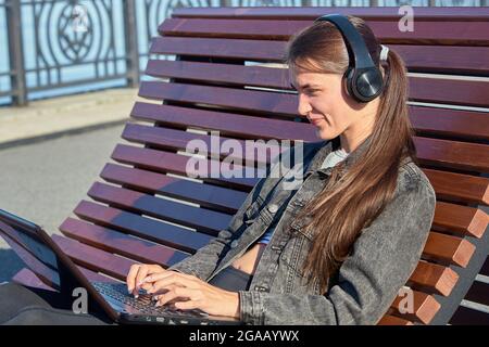 Eine Frau mit einem Laptop sitzt auf einer Außenbank und arbeitet online Stockfoto