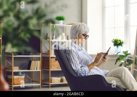 Seriöse ältere Frau, die im Wohnzimmer im Sessel sitzt und ihr Mobiltelefon benutzt Stockfoto