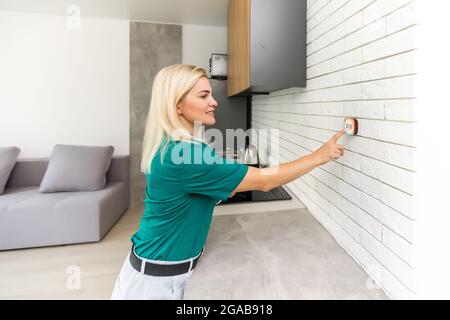 Frau, die die Heiztemperatur mit einem modernen, kabellosen Thermostat reguliert, der an der weißen Wand zu Hause installiert ist. Smart Home Heizung Regelkonzept Stockfoto