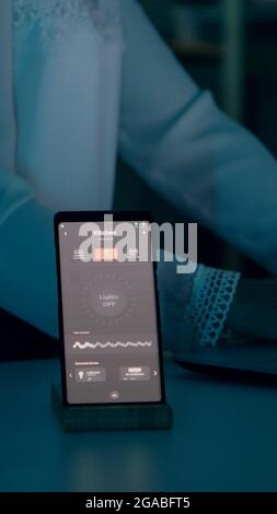 Frau tippt am Laptop, der zu Hause mit einem automatischen Beleuchtungssystem sitzt, das per Sprachsteuerung auf dem Smartphone die Glühbirnen anmacht. Lautsprecher-Gadget reagiert auf Befehle, Person, die das Umgebungslicht steuert Stockfoto