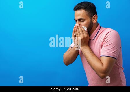 Junger Mann in rosa Hemd mit Allergie oder Kälte, bläst seine Nase in ein Gewebe vor blauem Hintergrund Stockfoto