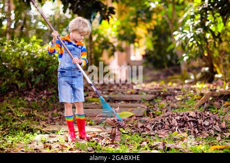 Kinder und Rechen im Herbstgarten. Kind beim Laub im Herbst. Gartenarbeit in der Laubsaison. Kleiner Junge hilft bei der Reinigung des Hinterhofs. Blattstapel auf Rasen. Stockfoto