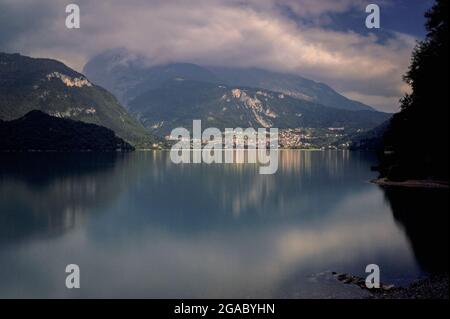 Das ruhige Trentino spiegelte sich in Molvenos nebliger Spiegelfassade … wider Die ruhige Oberfläche des Lago di Molveno, der als ‘der schönste See Italiens’ gilt, spiegelt die von einer dicken Wolke umhüllten Brenta-Dolomiten-Gipfel über dem kleinen Städtchen Molveno am nördlichen Ende des Sees in Trentino-Südtirol wider. Die rund 1,100 Einwohner von Molveno werden jeden Sommer von internationalen Besuchern angeschwollen, die von der Schönheit des alpinen Stausees, den Stränden, dem sauberen Wasser und den Aktivitäten wie Segeln, Windsurfen, Angeln, Kanufahren und Mountainbiken angezogen werden. Stockfoto