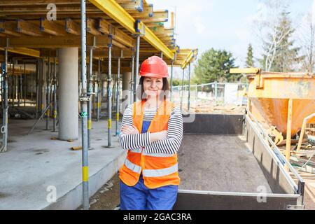 Zufriedene Frau mit Schutzhelm als Lehrling auf der Schale der Baustelle mit gefalteten Armen Stockfoto