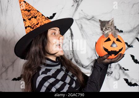 Eine Frau in einem Hexenkostüm in einem Hut hält einen Eimer-Kürbis mit Süßigkeiten und einen Ball. Halloween-Leute... Stockfoto