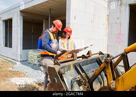 Architekt und Handwerker vor einem Bagger schauen sich den Bauplan auf einer Baustelle im Rohbau an Stockfoto