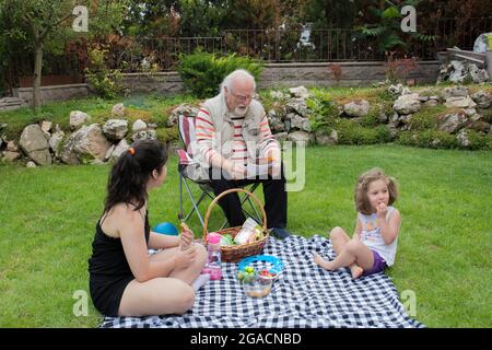 Die Mädchen picknicken mit ihren Großeltern. Der alte Mann sitzt auf dem Stuhl. Stockfoto