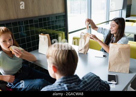 Fröhliches Teenager-Mädchen, das in der Schulkantine ein Sandwich in der Nähe von Klassenkameraden hält Stockfoto