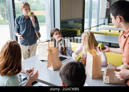 Fröhliche Teenager, die Sandwiches und Äpfel aßen, während sie sich im Speisesaal der Schule unterhielten Stockfoto