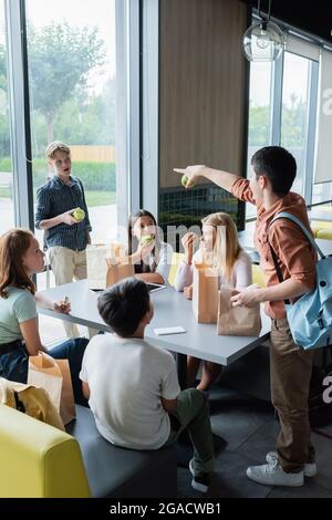 Teenager-Mädchen, das auf einen Klassenkameraden in der Nähe von Schülern zeigt, die im Speisesaal zu Mittag essen Stockfoto