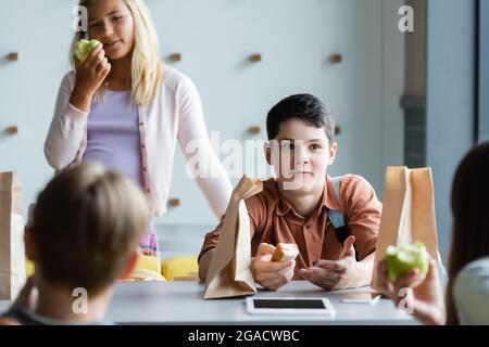 Erstaunter Teenager, der gestikuliert hat, während er mit verschwommenen Freunden in der Schulkantine sprach Stockfoto