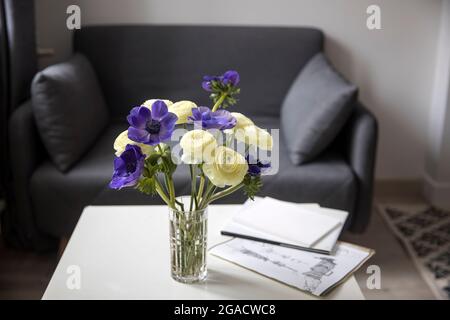 Bouquet von weißem Ranunculus und blauer Anemone in der Vase auf einem weißen Couchtisch neben dem Sofa mit karierten Rahmen. Die Bücher und die Zeitschrift. Schatten Stockfoto