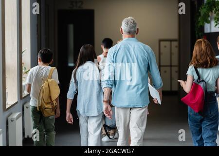 Rückansicht des Lehrers mit digitalem Tablet und der Schüler mit Rucksäcken, die durch den Schulkorridor laufen Stockfoto