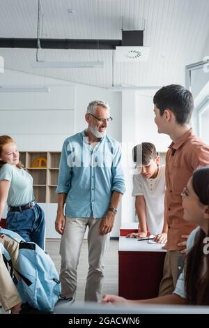 Reifer Lehrer lächelt in der Nähe multikultureller Teenager im Klassenzimmer Stockfoto