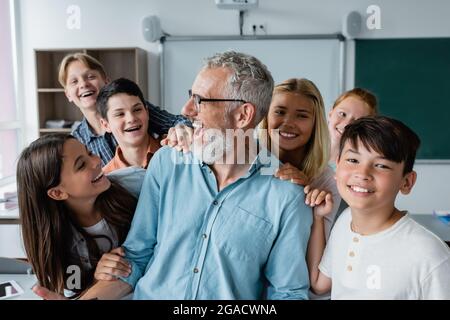 Glückliche multiethnische Schüler umarmen lachenden Lehrer im Klassenzimmer Stockfoto