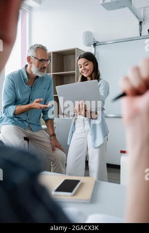 Fröhlicher Schüler, der seinen Laptop in der Nähe des Lehrers hält und mit der Hand zeigt Stockfoto