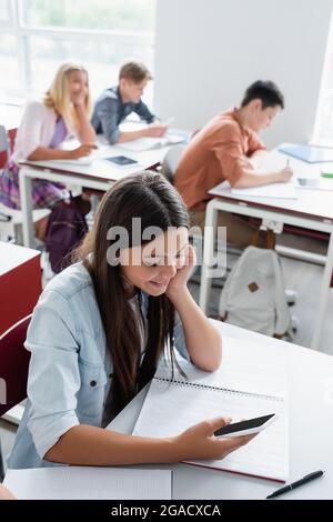 Lächelndes Schulmädchen mit Smartphone in der Nähe des Notebooks im Klassenzimmer Stockfoto