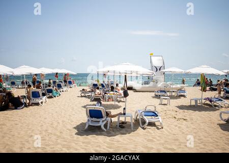 Sozopol, Burgas Bulgarien - 07 24 2021: Touristen liegen auf Sonnenliegen unter Sonnenschirmen, am Smokinya Strand. Ein Rettungsschwimmer mit einer gelben Flagge kann sein Stockfoto