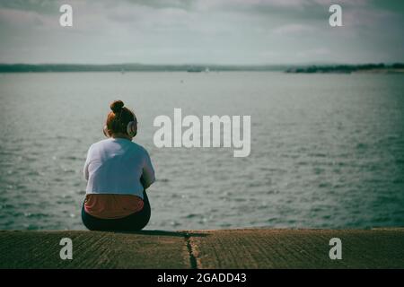 Ein Mädchen im Teenageralter, das Kopfhörer trägt, sitzt allein am Meer Stockfoto