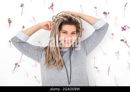 Fröhliche junge Frau in farbenfrohem Outfit, die mit einem Lächeln auf die Kamera blickt und Dreadlocks auf Blumenhintergrund schüttelt Stockfoto