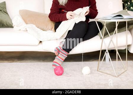 Zugeschnittenes Foto einer unbekannten Frau, die im Wohnzimmer auf der Couch sitzt und eine Strickdecke mit Mustern mit Nadeln strickt, und einer glücklichen Frau, die sich zu Hause entspannt, während sie es tut Stockfoto