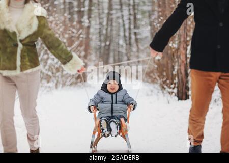 Junge schöne dreiköpfige Familie, die im verschneiten Wald spazieren geht, Mama und Papa schleppen ihren kleinen Sohn auf dem Schlitten und haben Spaß im Winterurlaub draußen. Saisonal geöffneter Urlaub Stockfoto