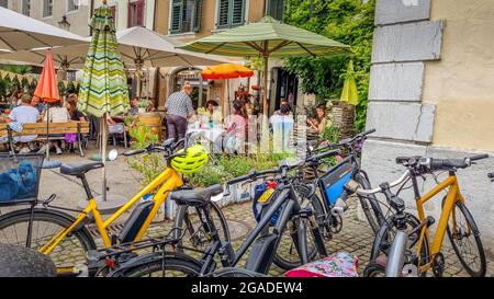 Gruppen sitzen am Tisch unter Sonnenschirmen im Freien, auf einem grünen Hof, in einer Straßenbar während der Aperitif-Stunde. Geparkte Elektrofahrräder mit Helm. Stockfoto